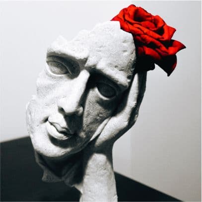 A white sculpted face resting on a hand with a single red rose behind it